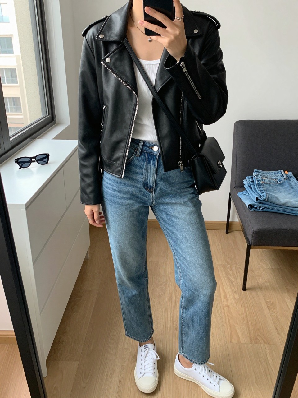 mirror selfie of a woman wearing the exact outfit described above, clearly showing a black faux leather moto jacket with silver hardware, fitted white ribbed tank, medium-wash straight-leg jeans, white low-top sneakers, and a black crossbody bag with a simple strap, holding a phone in front of a mirror, cropped at NECK so the face does not show, with a relaxed casual selfie pose, in a stylish city apartment, natural window light, including sunglasses placed on a dresser and folded denim jeans on a bench, latest iPhone photo quality.