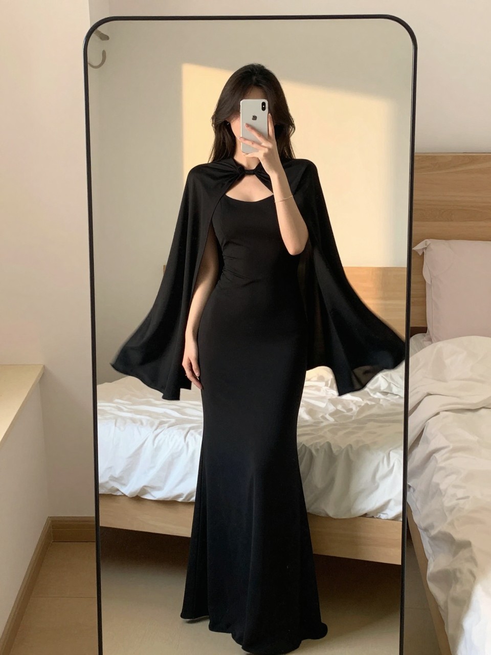 iPhone mirror selfie of a woman in a black maxi with a capelet, cape flowing behind, cropped at NECK so face doesn't show, in a feminine-themed bedroom, evening golden hour, iPhone photo quality.