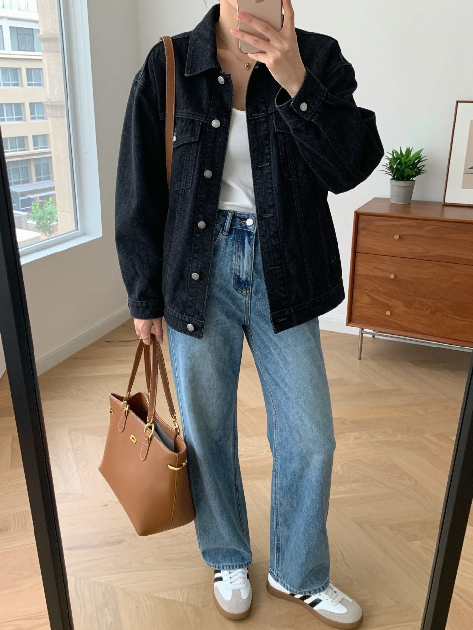 mirror selfie of a woman wearing the exact outfit described above, clearly showing a boxy-fit black denim jean jacket, white scoop-neck tank, blue high-waisted wide-leg jeans, white leather sneakers, and a tan shoulder bag with gold hardware, holding a phone in front of a mirror, cropped at NECK so the face does not show, with a relaxed casual selfie pose, in a stylish city apartment, natural window light, including a tote bag resting on the floor and a small plant on a dresser, latest iPhone photo quality.