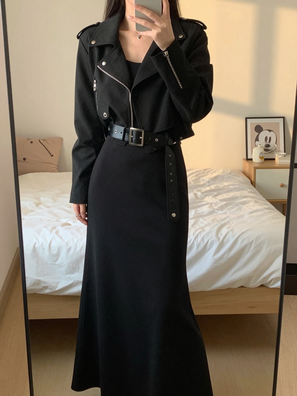 iPhone mirror selfie of a woman in a black maxi with a wide belt and moto jacket, posing from the side, cropped at NECK so face doesn't show, in a feminine-themed bedroom, evening golden hour, iPhone photo quality.