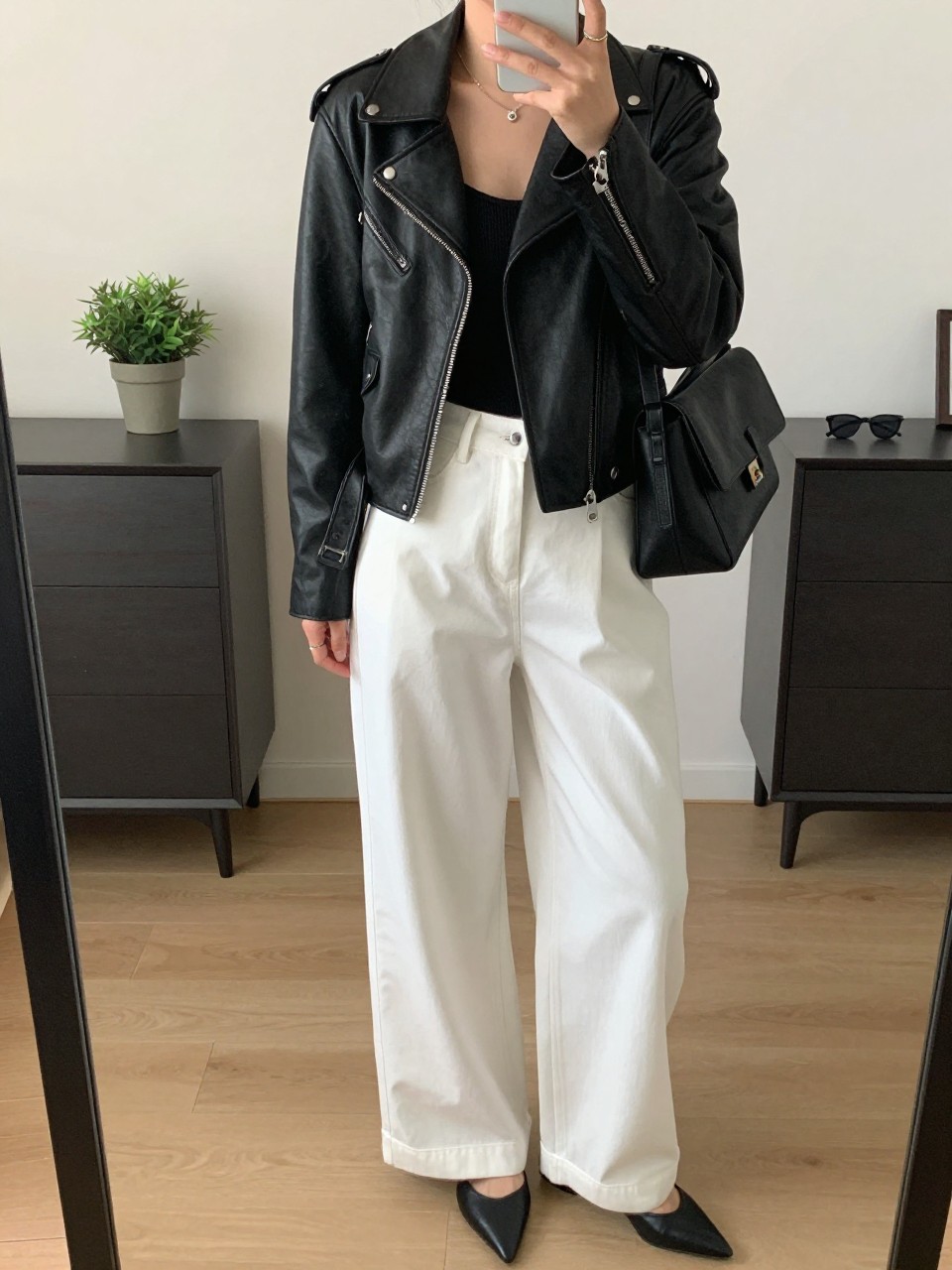 mirror selfie of a woman wearing the exact outfit described above, clearly showing a black leather biker jacket with silver hardware, soft black fitted bodysuit, white wide-leg trousers in smooth woven fabric, black pointed flats, and a black structured shoulder bag, holding a phone in front of a mirror, cropped at NECK so the face does not show, with a relaxed casual selfie pose, in a stylish city apartment, soft indoor lighting, including a small plant on a dresser and sunglasses placed on a dresser, latest iPhone photo quality.