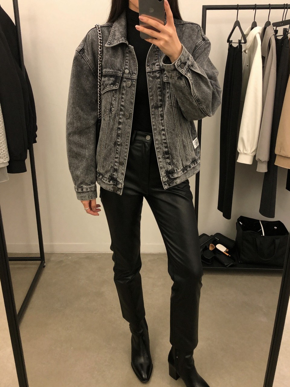 mirror selfie of a woman wearing the exact outfit described above, clearly showing a washed gray relaxed-fit denim jean jacket, black fitted bodysuit, black faux leather straight-leg pants, black ankle boots, and a silver chain-strap shoulder bag, holding a phone in front of a mirror, cropped at NECK so the face does not show, with a relaxed casual selfie pose, in a minimal dressing room, warm moody lighting, including a clothing rack with garments behind and a makeup brush or cosmetic bag on the counter, latest iPhone photo quality.