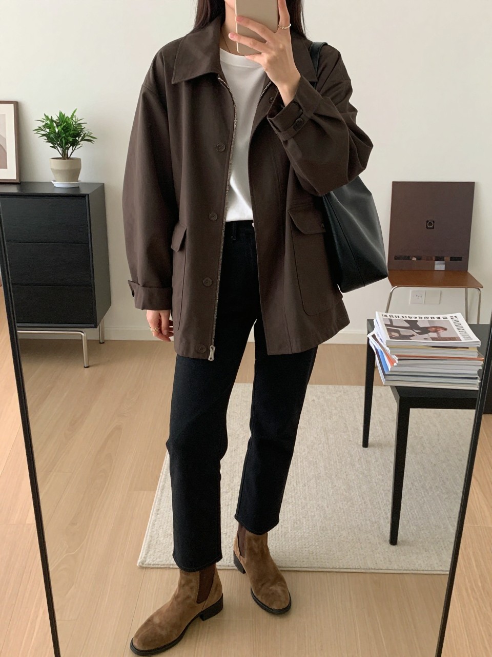 mirror selfie of a woman wearing the exact outfit described above, clearly showing a dark brown relaxed-shape barn jacket, white crew-neck soft cotton tee, black straight-leg jeans, brown suede ankle boots, and a black leather shoulder bag, holding a phone in front of a mirror, cropped at NECK so the face does not show, with a relaxed casual selfie pose, in a stylish city apartment, soft indoor lighting, including a small plant on a dresser and stack of fashion magazines on a side table, latest iPhone photo quality.