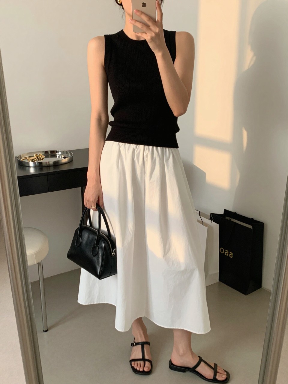 mirror selfie of a woman wearing the exact outfit described above, clearly showing a black sleeveless ribbed knit top, a white full cotton midi skirt, black leather strappy sandals, a black structured leather mini bag, and small gold stud earrings, holding a phone in front of a mirror, cropped at NECK so the face does not show, with a relaxed casual selfie pose, in a fashion studio mirror corner, evening golden hour, including jewelry tray on the vanity and shopping bags beside a chair, latest iPhone photo quality. (no watermarks on images)