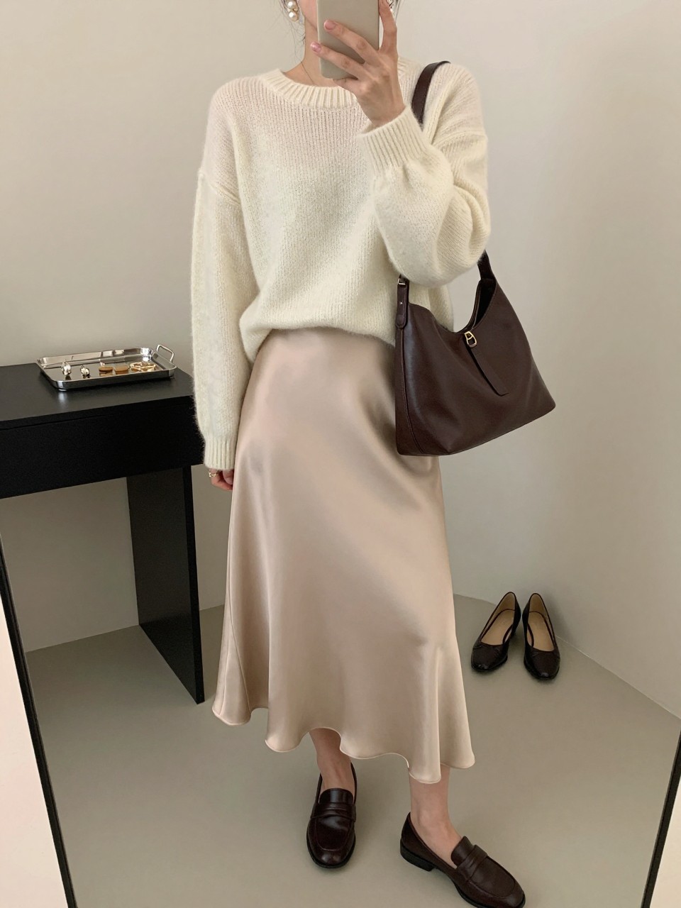mirror selfie of a woman wearing the exact outfit described above, clearly showing a soft cream lightweight knit sweater, a champagne satin midi skirt, dark brown leather loafers, a rich chocolate leather shoulder bag, and pearl drop earrings, holding a phone in front of a mirror, cropped at NECK so the face does not show, with a relaxed casual selfie pose, in a fashion studio mirror corner, soft indoor lighting, including jewelry tray on the vanity and pair of heels on the floor nearby, latest iPhone photo quality. (no watermarks on images)