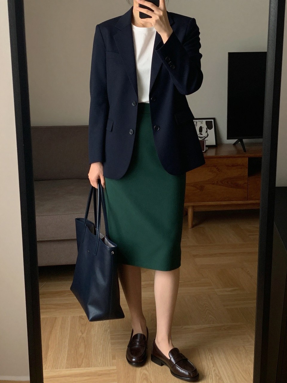 iPhone mirror selfie of a woman in a navy blazer over a white shell and hunter green pencil skirt with brown loafers, holding a navy tote, cropped at NECK so face doesn't show, in a city apartment, warm moody lighting, iPhone photo quality.