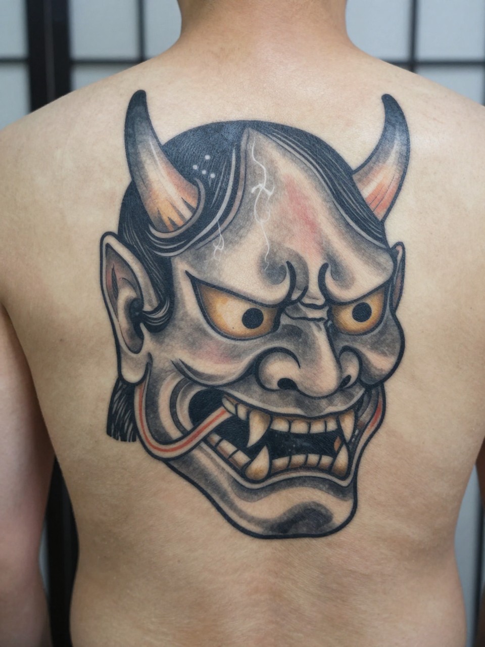 design-focused close-up of a full back Japanese Hannya mask tattoo on a man, fierce mask face, horns, sharp teeth, smoke filler, black and grey ink, strong traditional linework, soft shading, realistic skin texture, minimal blurred background (no watermarks on images)