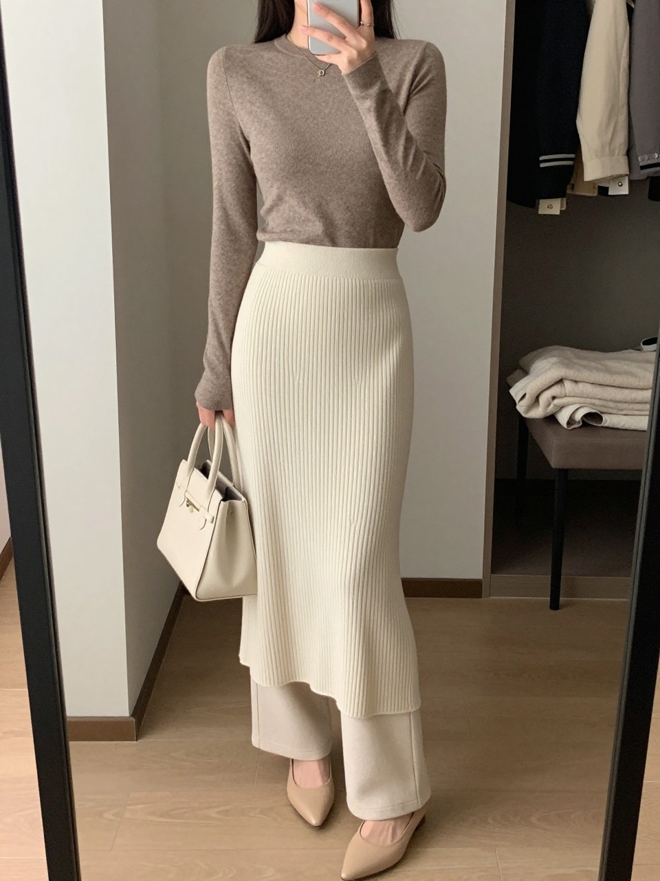 mirror selfie of a woman wearing a cream ribbed knit midi skirt layered over ivory straight-leg trousers, a taupe fitted long-sleeve knit top, beige pointed flats, and a cream structured handbag, holding a phone in front of a mirror, cropped at NECK so the face does not show, with a relaxed casual selfie pose, in a walk-in closet mirror, soft indoor lighting, including an open closet with clothes visible and a folded blanket on a chair, latest iPhone photo quality. (no watermarks on images)