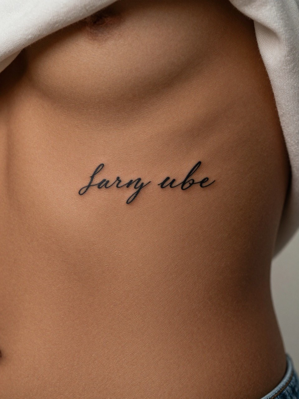 "extreme close-up of a small script word tattoo on deep dark skin at the ribcage, clean black ink, neat fine line lettering, tiny elegant style, realistic skin texture, soft fabric edge nearby, minimal background, design-focused feminine tattoo inspiration image (no watermarks on images)"