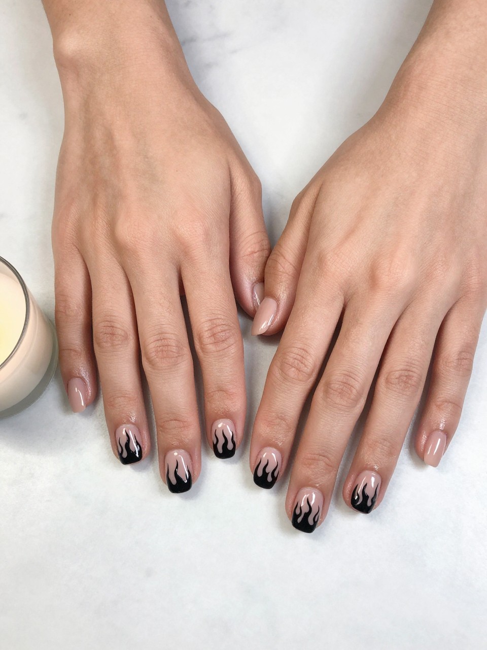 90-degree overhead top-down view of two feminine hands with rounded square medium nails.
The manicure features a soft nude base with black flame art painted across three accent nails.
Include glossy gel finish.

Hands are posed resting flat.

Scene set on a white marble surface with a small candle nearby.

Bright natural daylight with soft shadows, hyper-realistic lifestyle photography, sharp focus on nails, natural skin texture, realistic hands, no extra fingers.