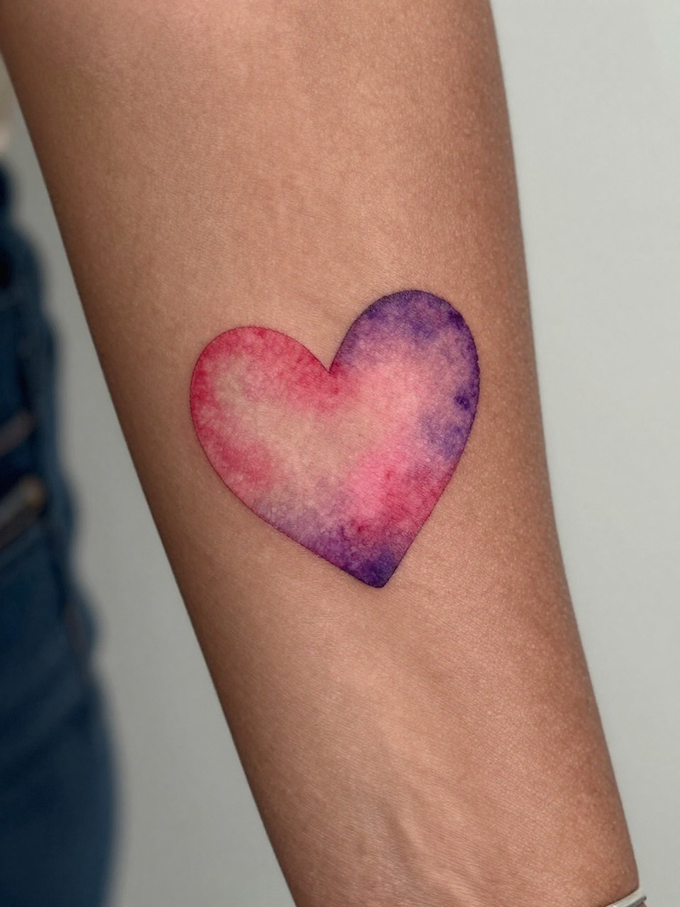 close-up of a watercolor-style heart tattoo on deep brown skin at the inner wrist, pink red and purple blended tones, soft edges, clean heart shape, realistic skin texture, delicate bracelet visible, design-focused close-up, blurred neutral background, modern tattoo photography (no watermarks on images)