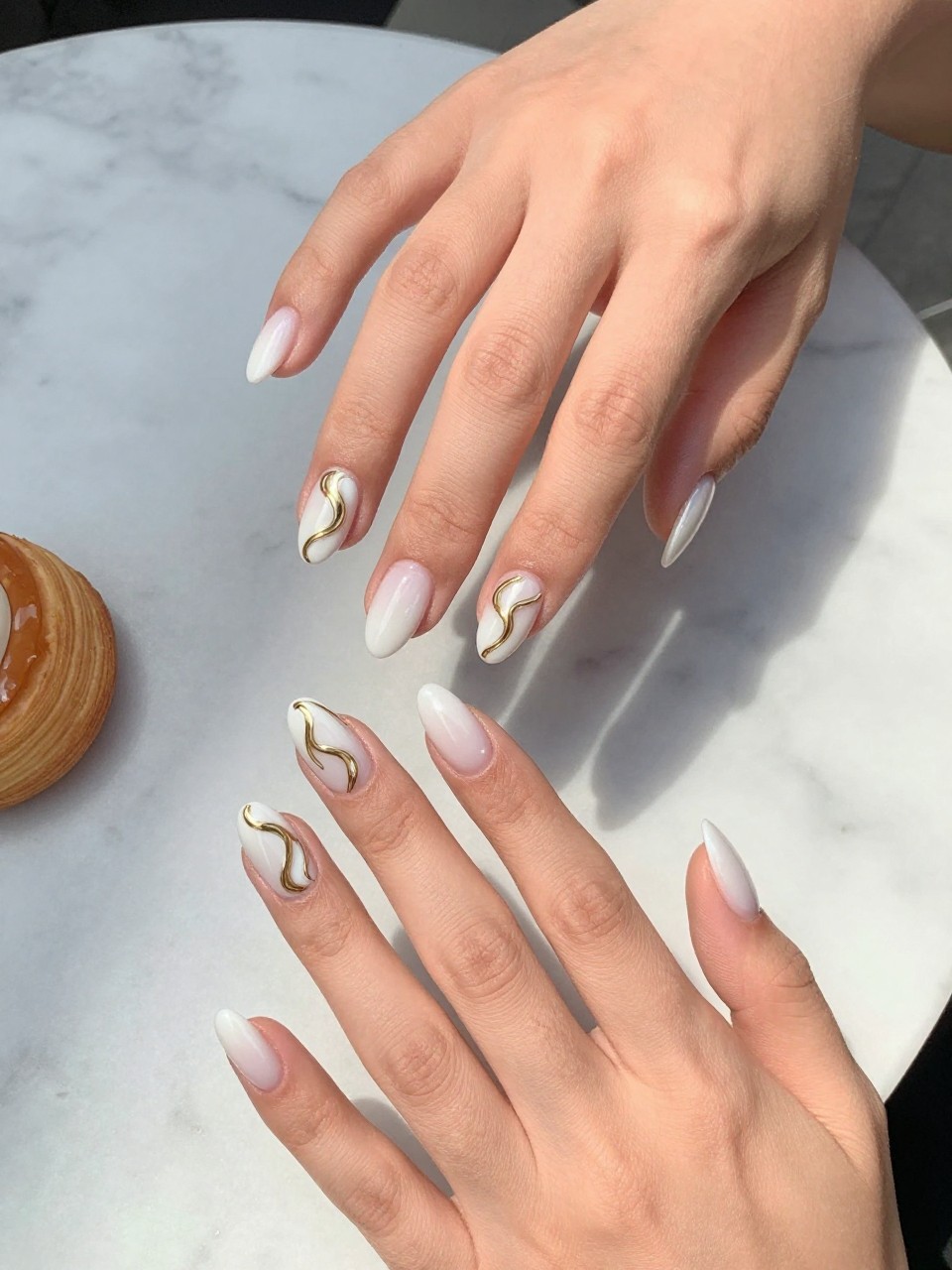 90-degree overhead top-down view of two feminine hands with almond medium nails.
The manicure features a milky white base with flowing gold chrome swirl designs across two accent nails.
Include reflective metallic accents with a glossy gel finish.

Hands are posed fingers gently spread.

Scene set on a marble café table with a small pastry nearby.

Bright natural daylight with soft shadows, hyper-realistic lifestyle photography, sharp focus on nails, natural skin texture, realistic hands, no extra fingers.