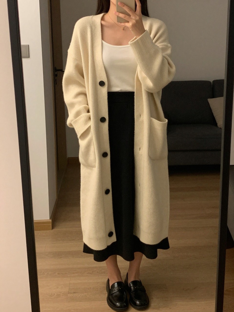 iPhone mirror selfie of a woman in a long cream cardigan over a tank and black skirt with loafers, standing with hands in cardigan pockets, cropped at NECK so face doesn't show, in a city apartment, warm moody lighting, iPhone photo quality.