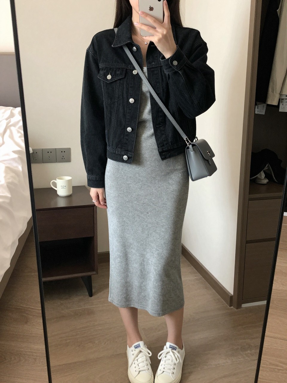 mirror selfie of a woman wearing the exact outfit described above, clearly showing a fitted black denim jean jacket with a clean shape, heather gray knit midi dress in soft stretch fabric, white low-top sneakers, gray crossbody bag with silver hardware, and simple silver stud earrings, holding a phone in front of a mirror, cropped at NECK so the face does not show, with a relaxed casual selfie pose, in a cozy bedroom mirror setup, soft indoor lighting, including a coffee mug on a bedside table and an open closet with clothes visible, latest iPhone photo quality.