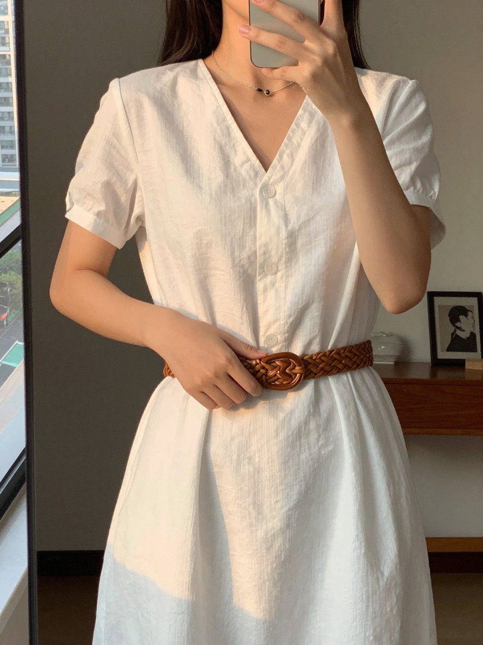 iPhone mirror selfie of a woman in a white linen dress with a woven belt, adjusting the belt, cropped at the neck so face doesn't show, in a city apartment, evening golden hour lighting, iPhone photo quality.
