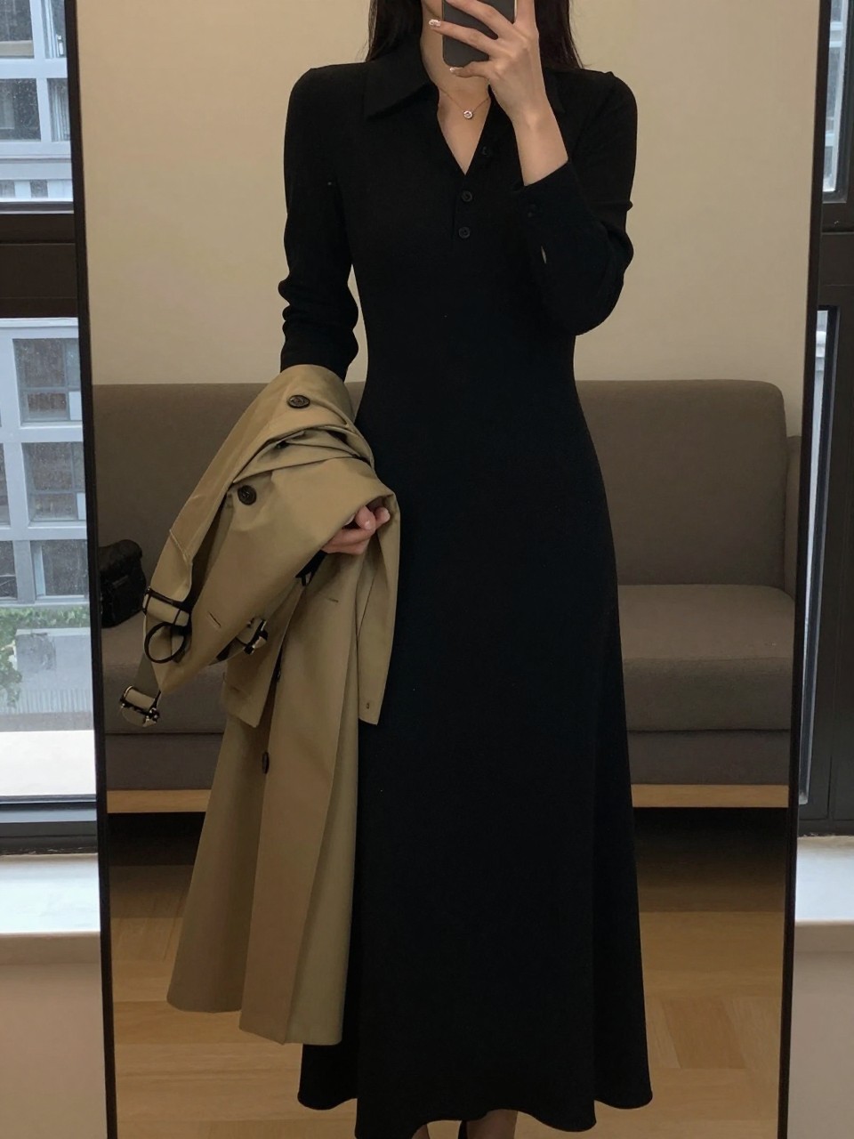 iPhone mirror selfie of a woman in a collared black maxi dress, holding a trench coat, cropped at NECK so face doesn't show, in a city apartment, warm moody lighting, iPhone photo quality.
