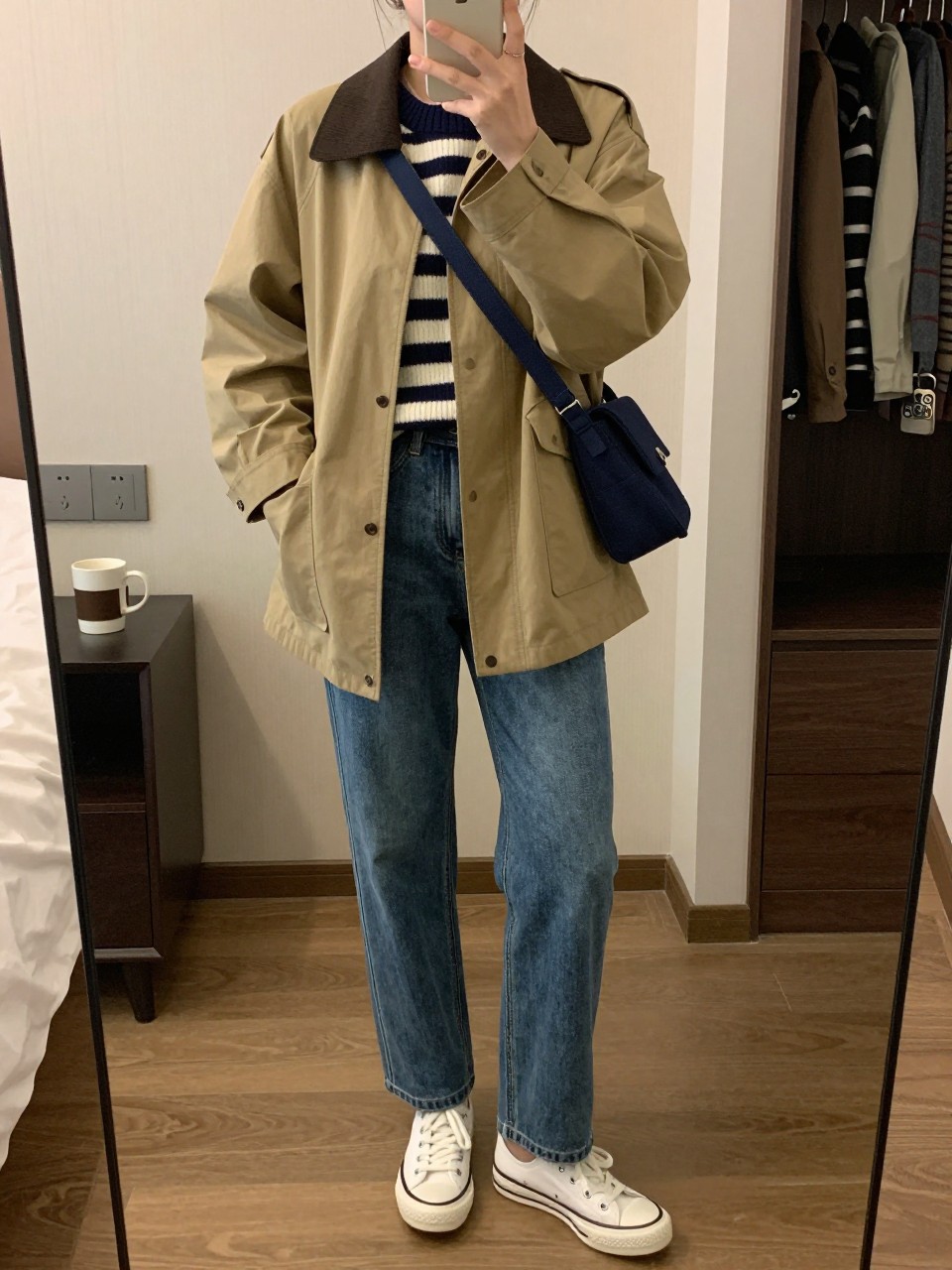 mirror selfie of a woman wearing the exact outfit described above, clearly showing a khaki barn jacket with dark brown trim, navy and cream striped knit sweater, blue straight-leg jeans, white canvas sneakers, and a navy crossbody bag, holding a phone in front of a mirror, cropped at NECK so the face does not show, with a relaxed casual selfie pose, in a cozy bedroom mirror setup, soft indoor lighting, including a coffee mug on a bedside table and open closet with clothes visible, latest iPhone photo quality.