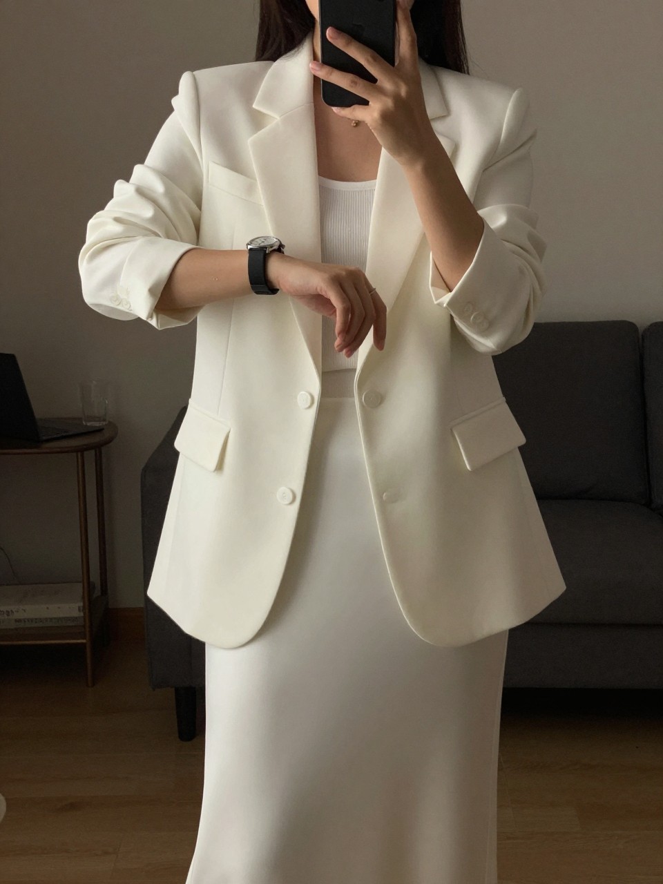 iPhone mirror selfie of a woman in a long-sleeve maxi with a white blazer, checking her watch, cropped at NECK so face doesn't show, in a city apartment, warm moody lighting, iPhone photo quality.
