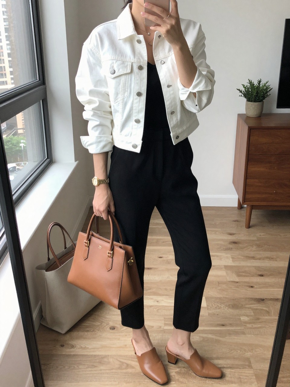 mirror selfie of a woman wearing the exact outfit described above, clearly showing a white denim jean jacket with a neat cropped fit, black sleeveless jumpsuit with tapered legs, tan heeled mules, cognac leather handbag, and a gold watch with simple studs, holding a phone in front of a mirror, cropped at NECK so the face does not show, with a relaxed casual selfie pose, in a stylish city apartment, natural window light, including a tote bag resting on the floor and a small plant on a dresser, latest iPhone photo quality.