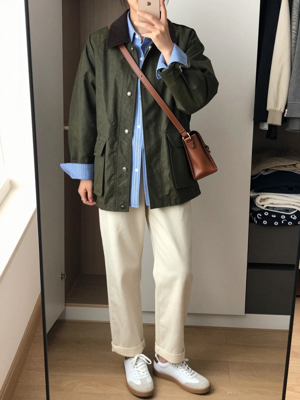 mirror selfie of a woman wearing the exact outfit described above, clearly showing an olive green waxed barn jacket with a dark collar, blue and white striped oversized shirt, cream straight-leg trousers, white leather sneakers, and a brown leather crossbody bag, holding a phone in front of a mirror, cropped at NECK so the face does not show, with a relaxed casual selfie pose, in a walk-in closet mirror, natural window light, including an open closet with clothes visible and folded blanket on a chair, latest iPhone photo quality.