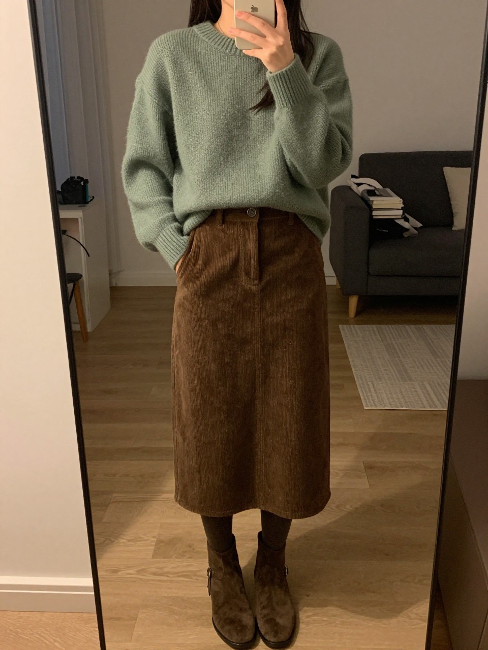 iPhone mirror selfie of a woman in a sage green sweater and brown corduroy skirt with tights and ankle boots, standing with hands in pockets, cropped at NECK so face doesn't show, in a city apartment, warm moody lighting, iPhone photo quality.