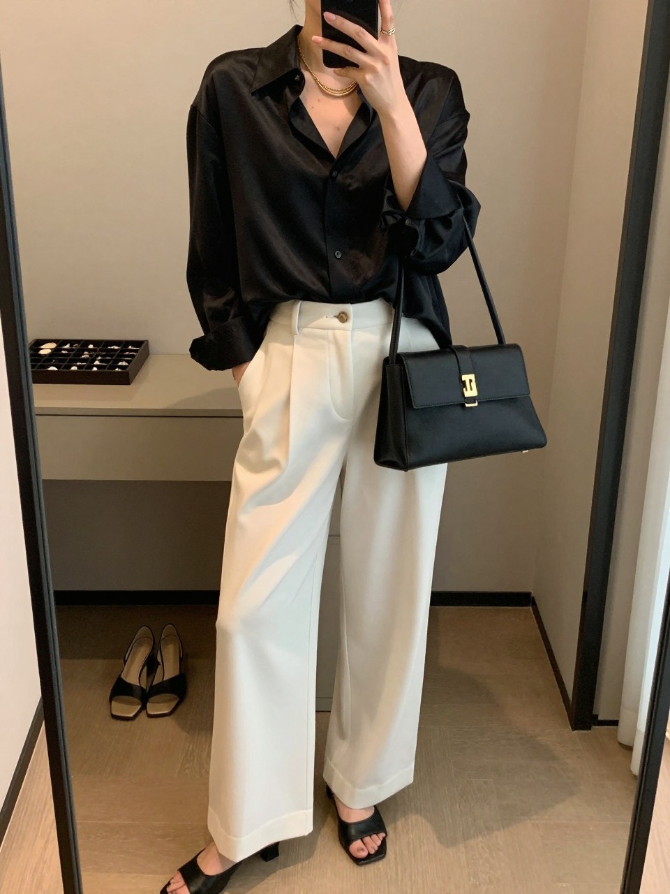 mirror selfie of a woman wearing a black oversized silk shirt with a soft sheen, white high-waisted wide-leg trousers, black heeled mules, a black structured mini bag, and a gold layered necklace, holding a phone in front of a mirror, cropped at NECK so the face does not show, with a relaxed casual selfie pose, in a minimal dressing room, warm moody lighting, including a jewelry tray on the vanity and a pair of heels on the floor nearby, latest iPhone photo quality. (no watermarks on images)