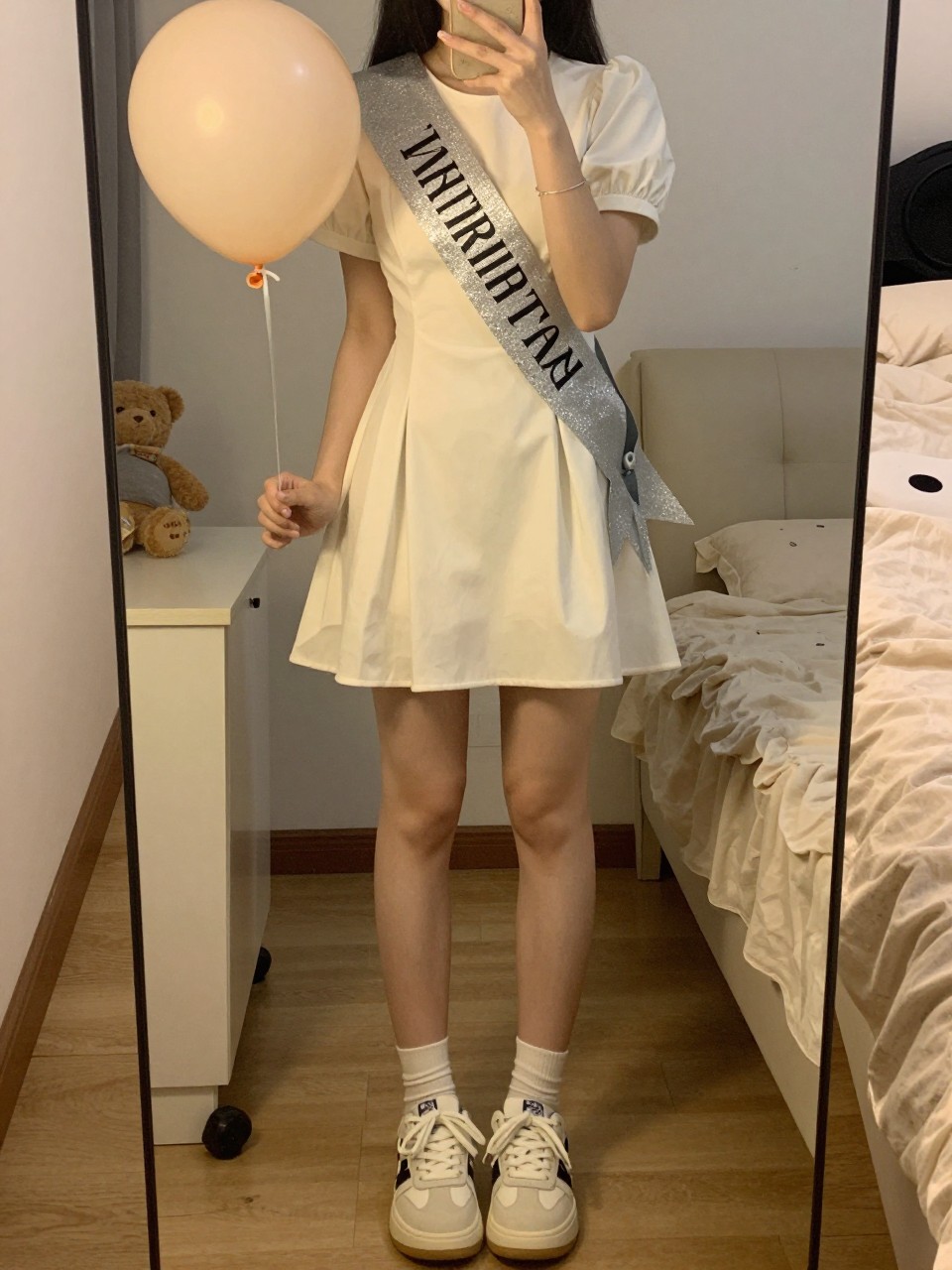 iPhone mirror selfie of a woman in a simple dress with a birthday sash and statement sneakers, holding a balloon, cropped at the neck so face doesn't show, in a feminine-themed bedroom, warm moody lighting, iPhone photo quality.