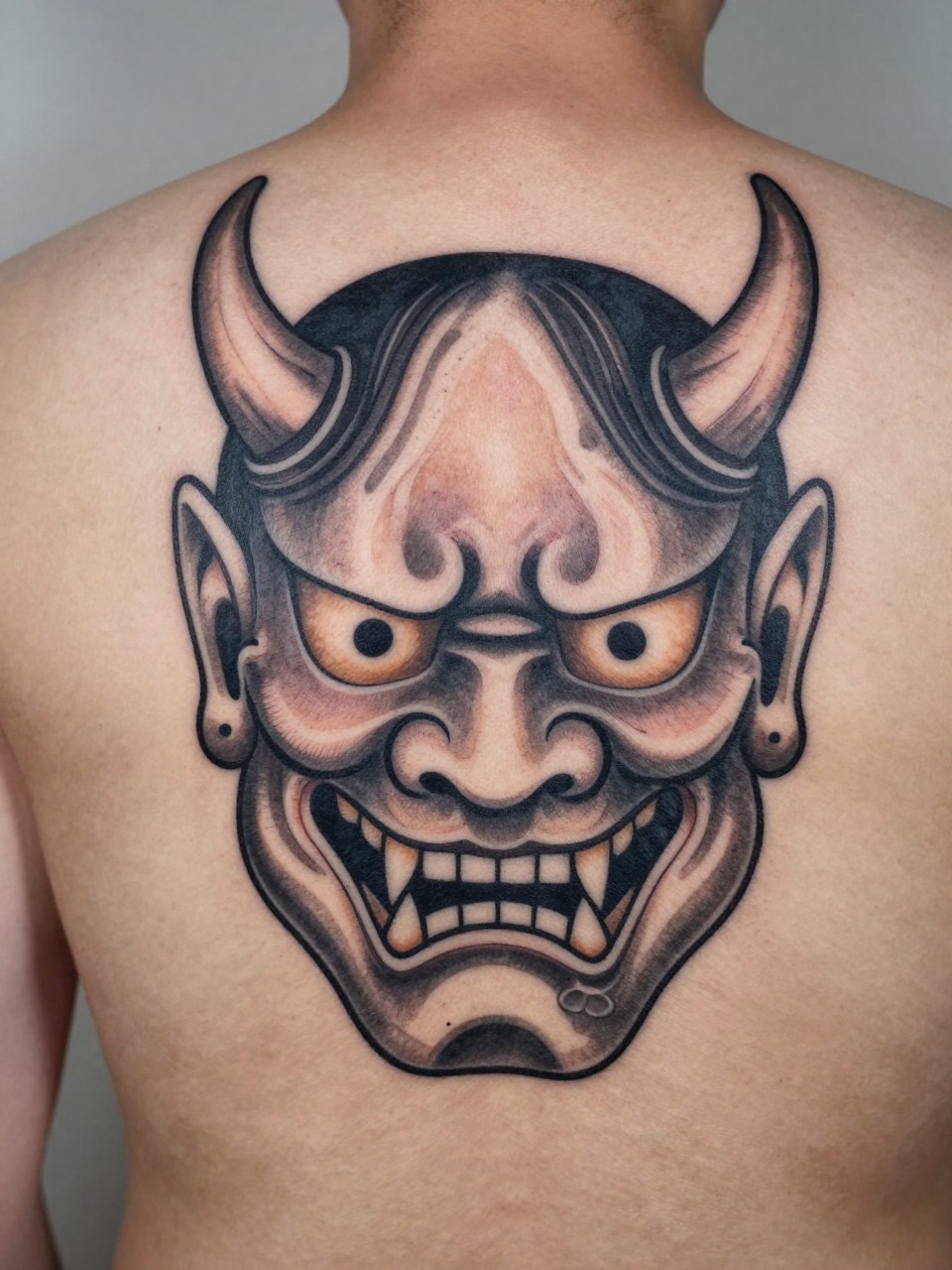 "macro shot of a full back Japanese Oni mask tattoo on a man, bold demon face, curved horns, flame details, black and grey ink, thick traditional linework, dramatic shading, realistic skin texture, design-first back tattoo composition, soft neutral background (no watermarks on images)"