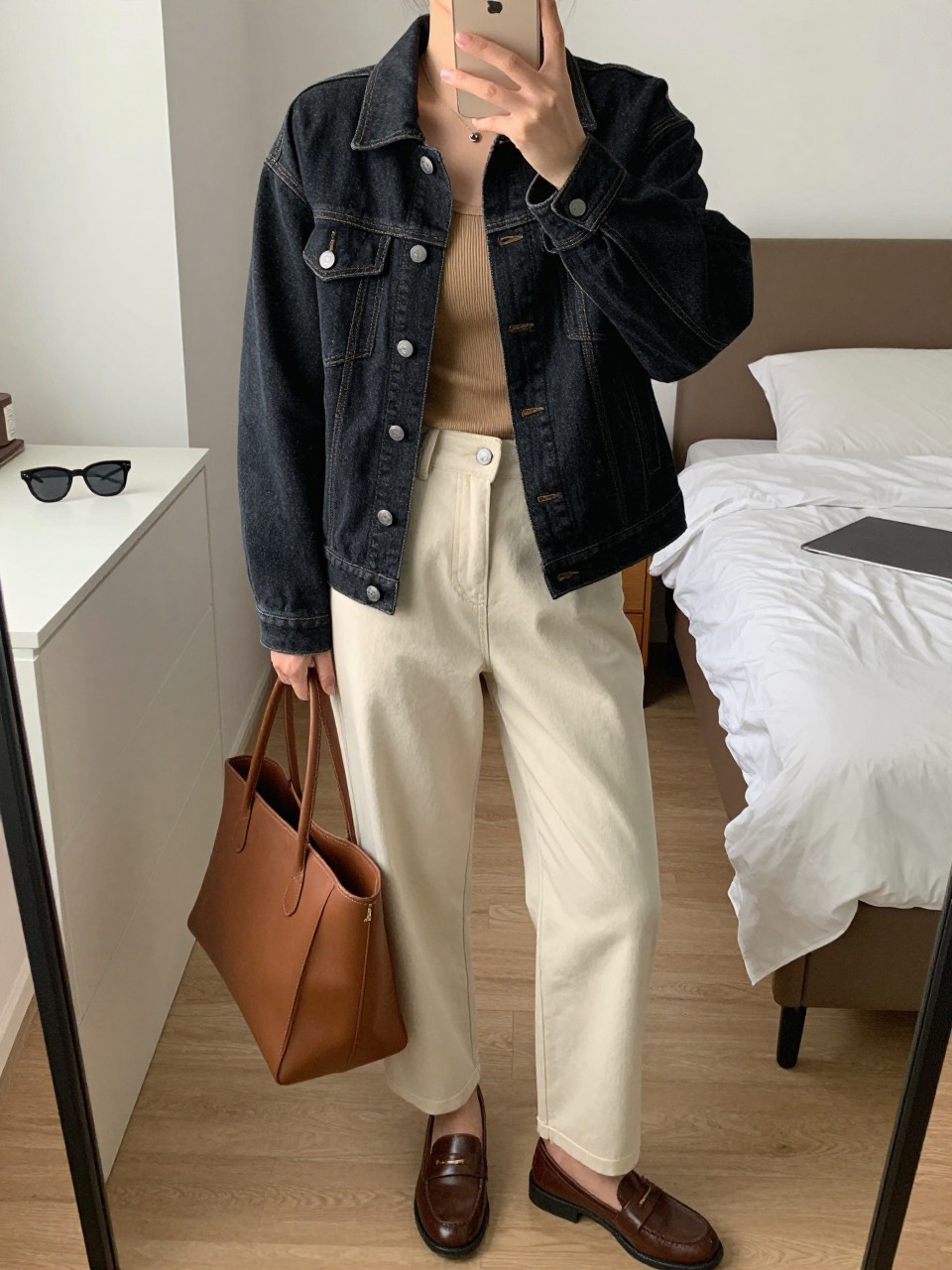mirror selfie of a woman wearing the exact outfit described above, clearly showing a black denim jean jacket with silver button details, camel fitted ribbed tank, cream straight-leg trousers, brown leather loafers, and a brown structured tote bag, holding a phone in front of a mirror, cropped at NECK so the face does not show, with a relaxed casual selfie pose, in a stylish city apartment, soft indoor lighting, including sunglasses placed on a dresser and a laptop or tablet on the bed, latest iPhone photo quality.