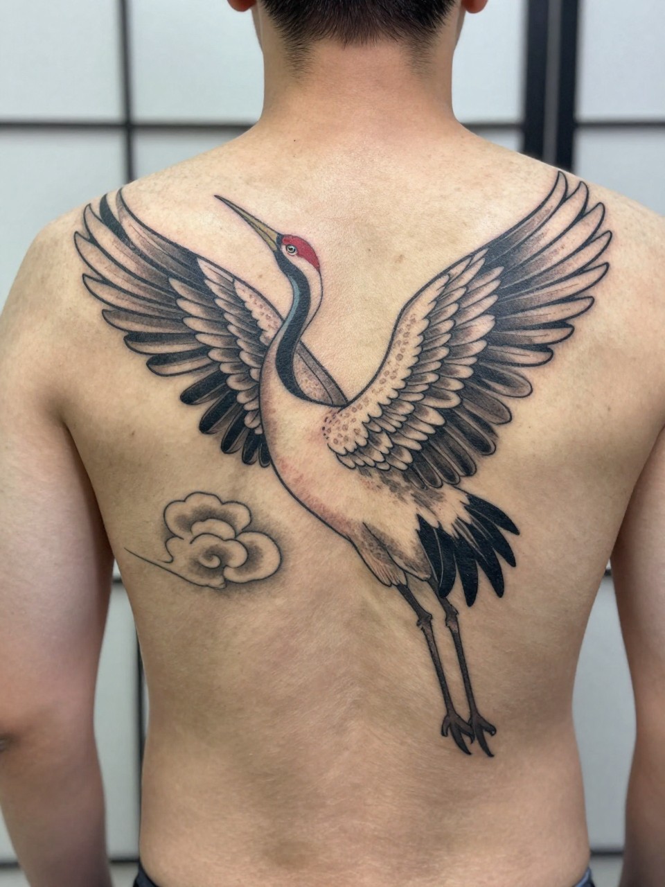 "design-focused close-up of a full back Japanese crane tattoo on a man, elegant crane with spread wings, cloud details across the back, black and grey ink, clean traditional linework, soft shading, realistic skin texture, minimal blurred background (no watermarks on images)"