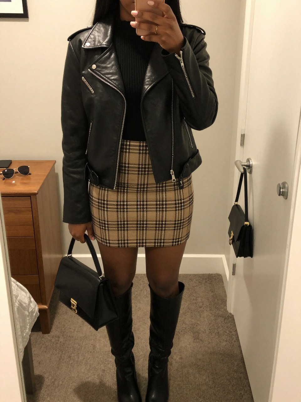 mirror selfie of a black woman wearing the exact outfit described above, clearly showing a black moto leather jacket with silver zip details, black fine-knit mock-neck top, brown and cream plaid mini skirt, black knee-high boots, and a structured black mini handbag, holding a phone in front of a mirror, cropped at NECK so the face does not show, with a relaxed casual selfie pose, in a feminine-themed bedroom, warm moody lighting, including sunglasses placed on a dresser and a handbag hanging on a door handle, latest iPhone photo quality.
