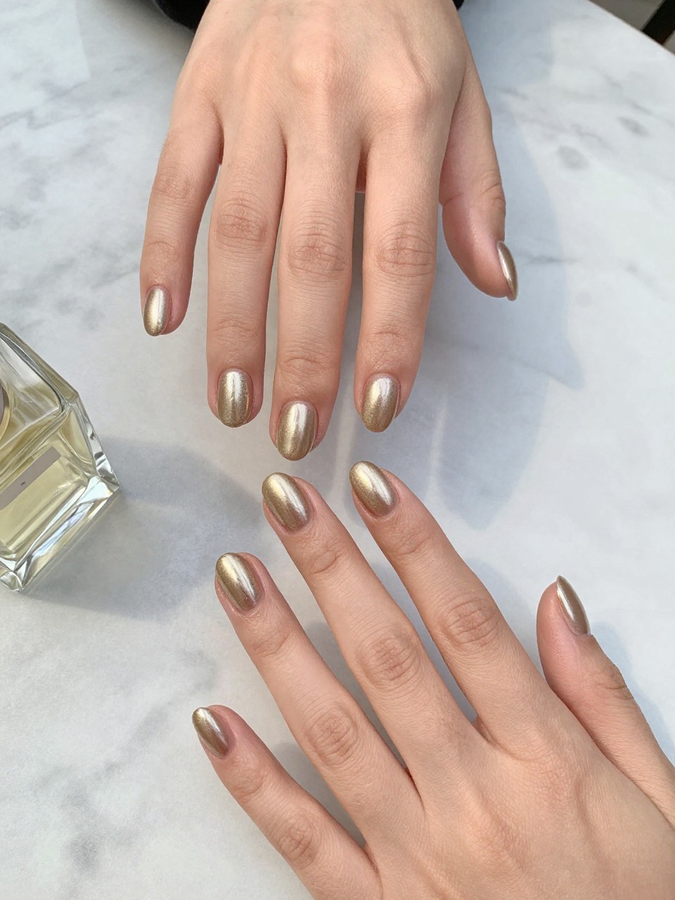 90-degree overhead top-down view of two feminine hands with oval medium nails.
The manicure features a champagne gold base on every nail with a soft metallic shimmer finish.
Include smooth reflective shine with a glossy gel finish.

Hands are posed fingers gently spread.

Scene set on a marble café table with a small glass perfume bottle nearby.

Bright natural daylight with soft shadows, hyper-realistic lifestyle photography, sharp focus on nails, natural skin texture, realistic hands, no extra fingers.