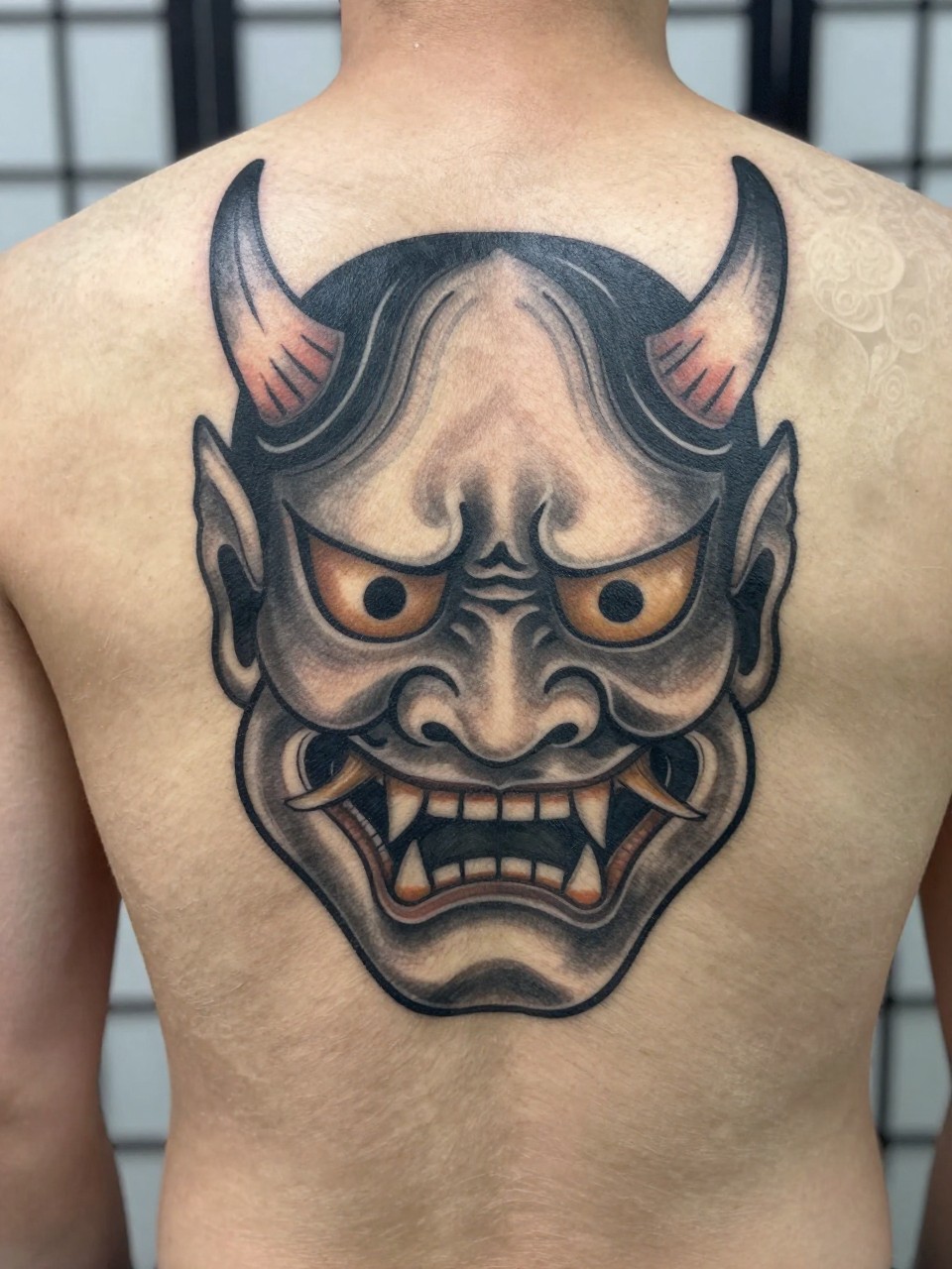 "design-focused close-up of a full back Japanese Hannya mask tattoo on a man, fierce mask face, horns, sharp teeth, smoke filler, black and grey ink, strong traditional linework, soft shading, realistic skin texture, minimal blurred background (no watermarks on images)"