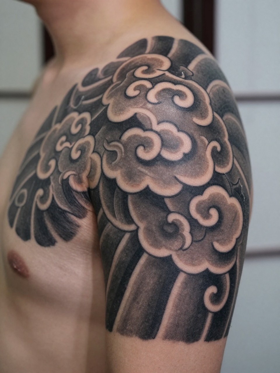 design-focused close-up of a Japanese cloud shoulder cap tattoo, bold traditional cloud forms shaped around the shoulder, black and grey ink, crisp linework, soft shading, realistic skin texture, minimal blurred background, modern tattoo inspiration photo (no watermarks on images)