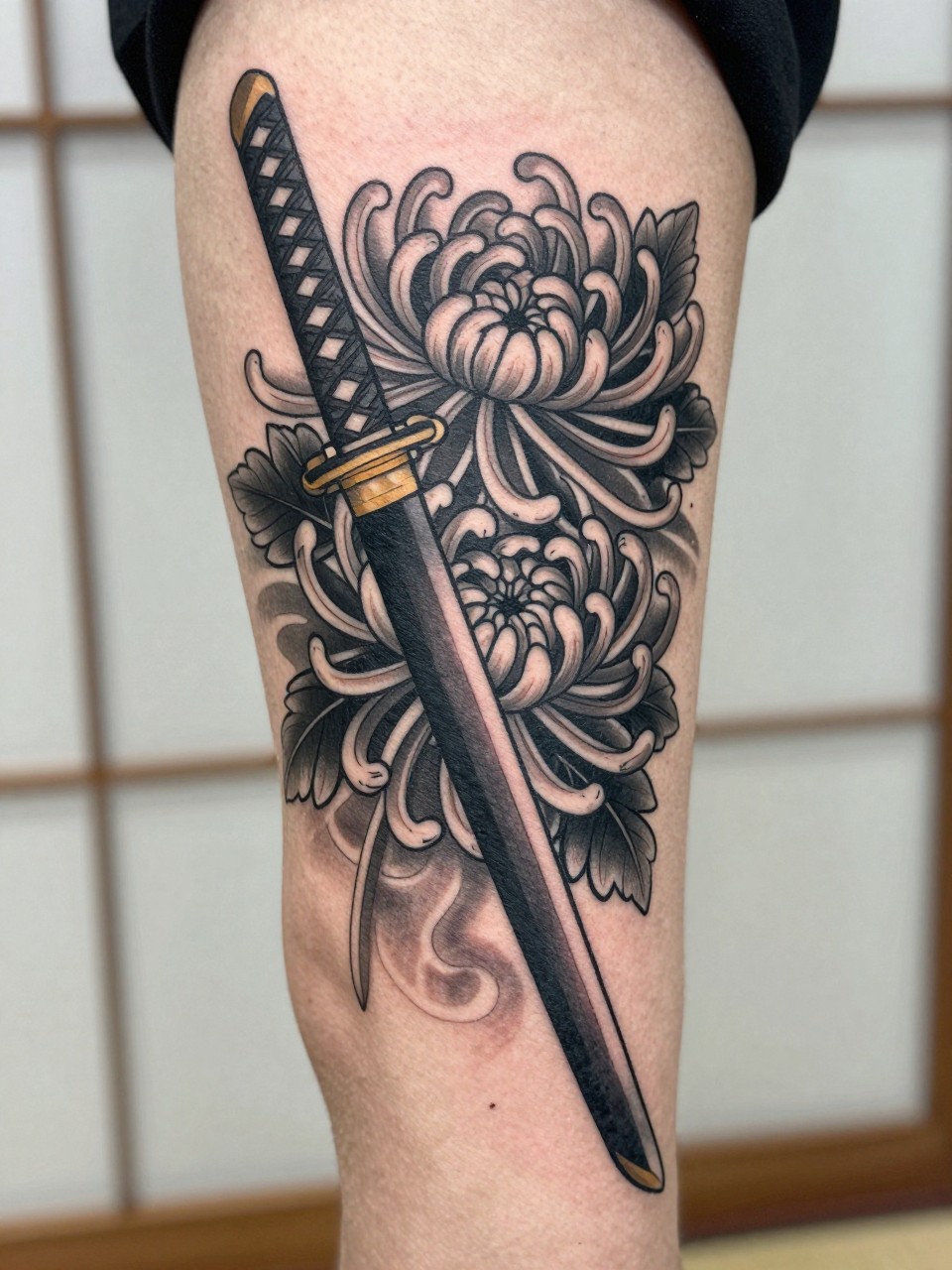 design-focused close-up of a traditional Japanese sword and chrysanthemum tattoo on the arm or thigh, katana detail crossing large chrysanthemum blooms, black and grey ink, clean bold linework, soft shading, realistic skin texture, minimal blurred background, modern tattoo inspiration photo (no watermarks on images)