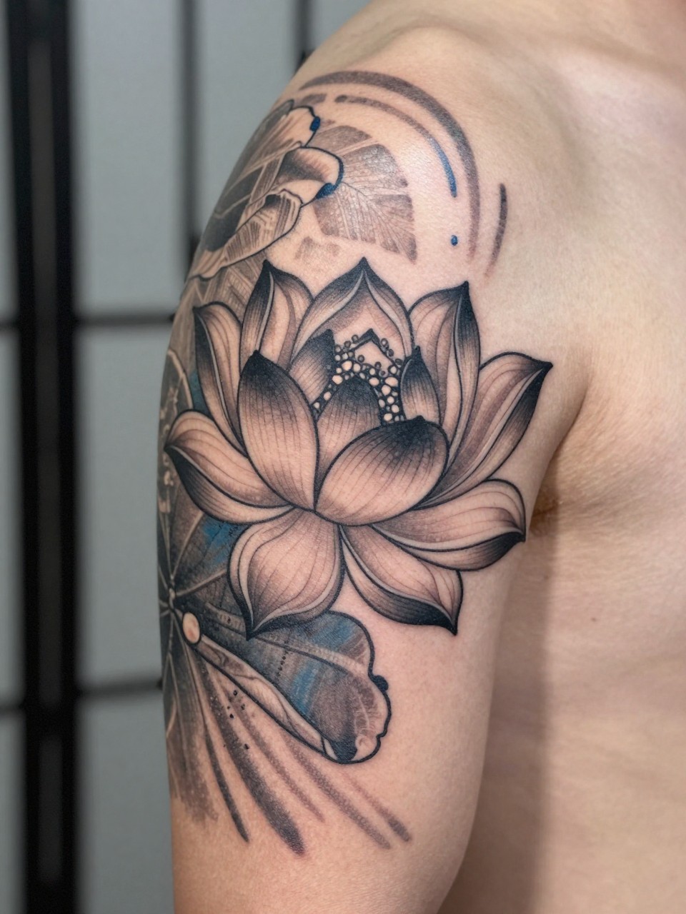 close-up of a Japanese lotus and flowing water tattoo on the upper arm, detailed lotus petals, black and grey ink, soft blue-grey shading in water accents, clean traditional linework, realistic skin texture, design-focused close-up, blurred neutral background, modern tattoo photography (no watermarks on images)