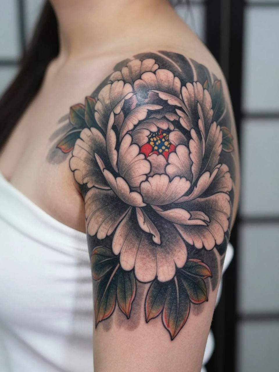 design-focused close-up of a bold Japanese peony tattoo on the shoulder, large layered petals, black and grey ink with subtle red accents, strong traditional linework, soft shading, realistic skin texture, off-shoulder top visible, minimal blurred background, modern tattoo inspiration photo (no watermarks on images)