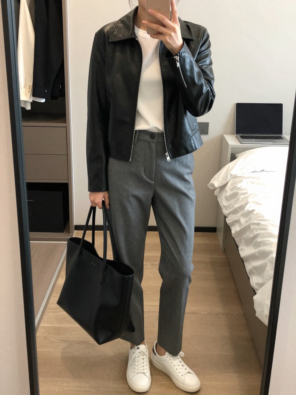 mirror selfie of a woman wearing the exact outfit described above, clearly showing a black minimal zip-front leather jacket, white crew-neck fitted tee, gray straight-leg tailored trousers, white leather sneakers, and a black tote bag with a clean shape, holding a phone in front of a mirror, cropped at NECK so the face does not show, with a relaxed casual selfie pose, in a walk-in closet mirror, soft indoor lighting, including an open closet with clothes visible and a laptop or tablet on the bed, latest iPhone photo quality.
