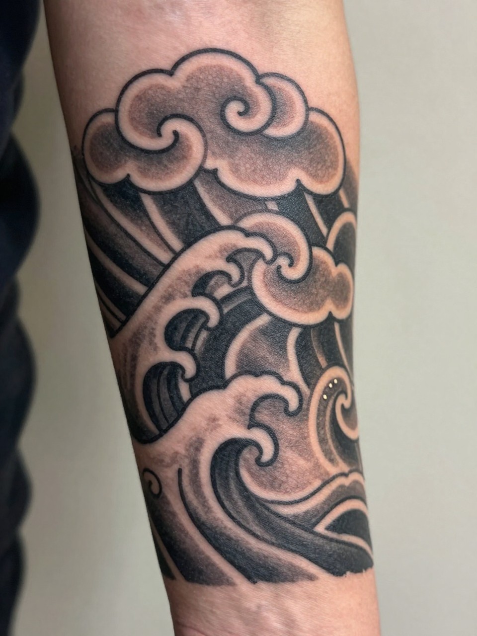 macro shot of a Japanese cloud and wave tattoo on the forearm, traditional cloud shapes above curling waves, black and grey ink, bold linework, smooth shading, realistic skin texture, design-first tattoo image, soft neutral background, tattoo inspiration image (no watermarks on images)