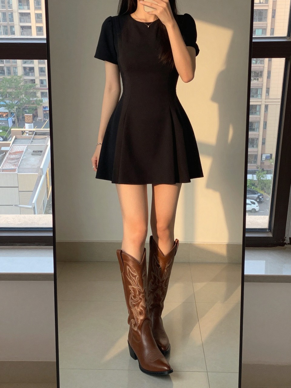 iPhone mirror selfie of a woman in a little black dress and statement cowboy boots, one foot slightly forward to show the boots, cropped at the neck so face doesn't show, in a city apartment, evening golden hour lighting, iPhone photo quality.