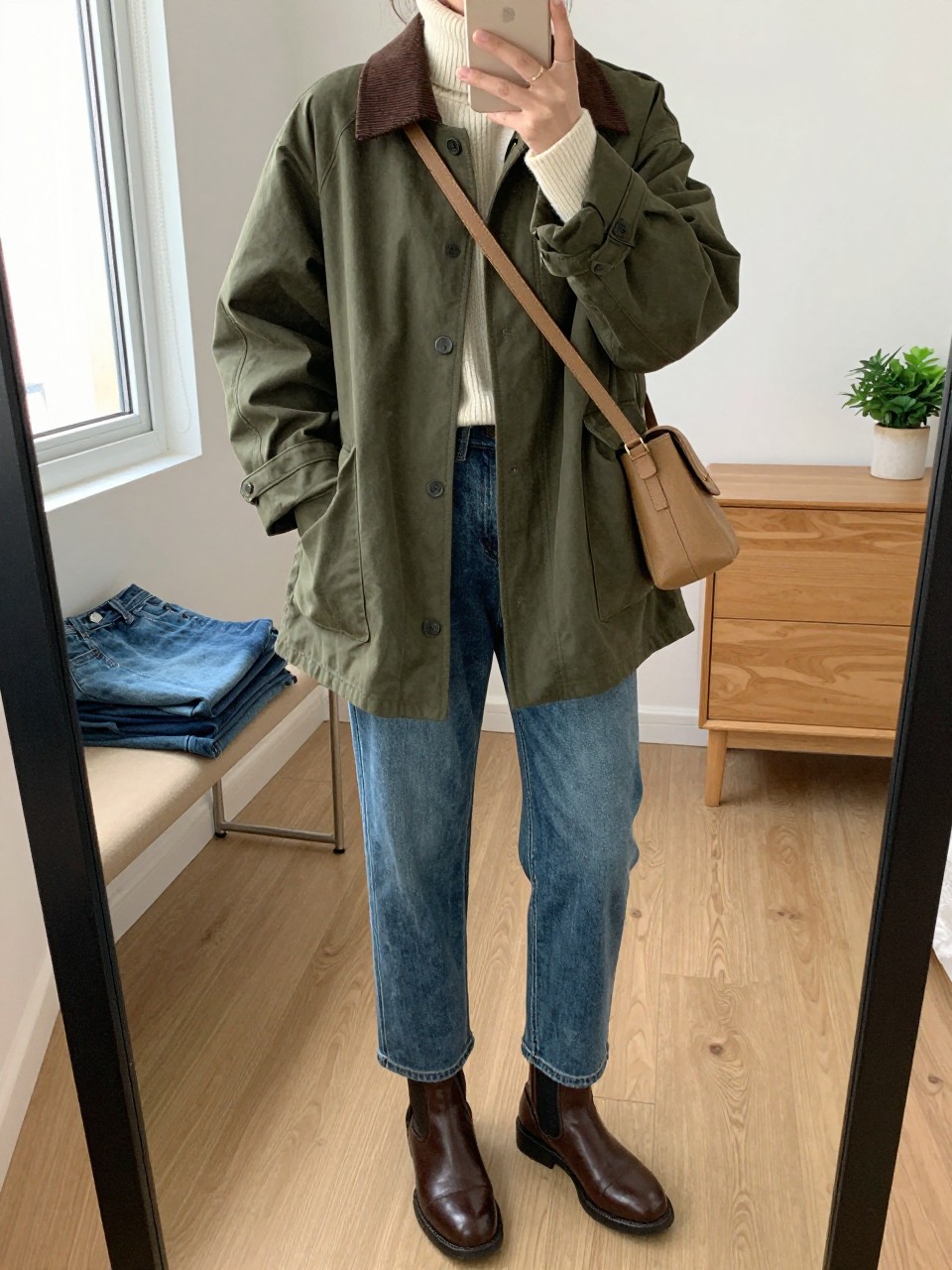mirror selfie of a woman wearing the exact outfit described above, clearly showing an olive cotton barn jacket with a brown corduroy collar, cream ribbed turtleneck sweater, medium-wash straight-leg jeans, brown leather ankle boots, and a tan crossbody bag with a simple strap, holding a phone in front of a mirror, cropped at NECK so the face does not show, with a relaxed casual selfie pose, in a cozy bedroom mirror setup, natural window light, including folded denim jeans on a bench and a small plant on a dresser, latest iPhone photo quality.