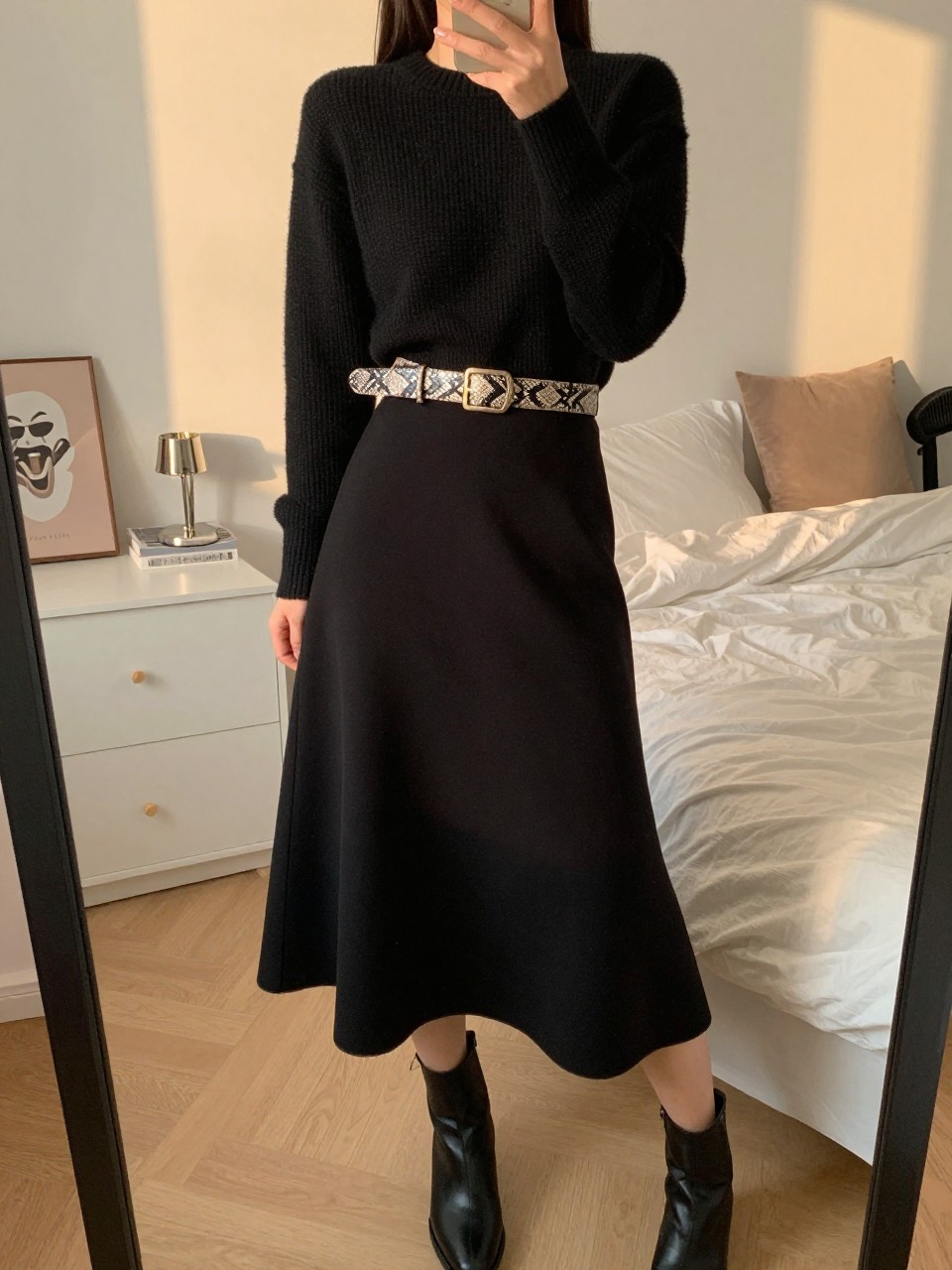 iPhone mirror selfie of a woman in a cropped black sweater and black full midi skirt with a wide snakeskin belt and ankle boots, one hand on belt, cropped at NECK so face doesn't show, in a feminine-themed bedroom, evening golden hour, iPhone photo quality.
