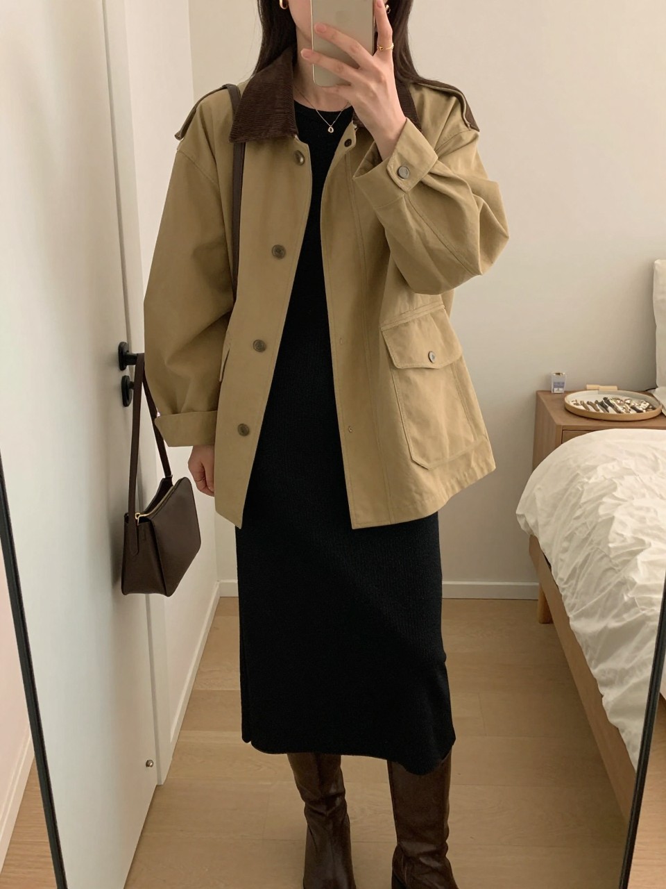 mirror selfie of a woman wearing the exact outfit described above, clearly showing a tan canvas barn jacket with a dark brown corduroy collar, black knit midi dress in soft stretch fabric, brown knee-high boots, dark brown shoulder bag, and small gold hoop earrings, holding a phone in front of a mirror, cropped at NECK so the face does not show, with a relaxed casual selfie pose, in a feminine-themed bedroom, soft indoor lighting, including a handbag hanging on a door handle and a jewelry tray on the vanity, latest iPhone photo quality.