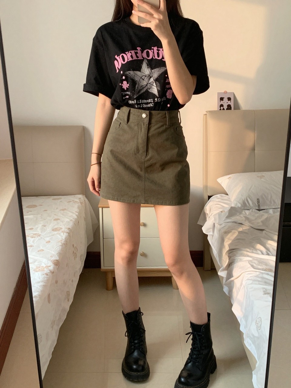 iPhone mirror selfie of a woman in a black graphic tee tucked into an olive green mini skirt with combat boots, standing with feet apart, cropped at NECK so face doesn't show, in a feminine-themed bedroom, evening golden hour, iPhone photo quality.