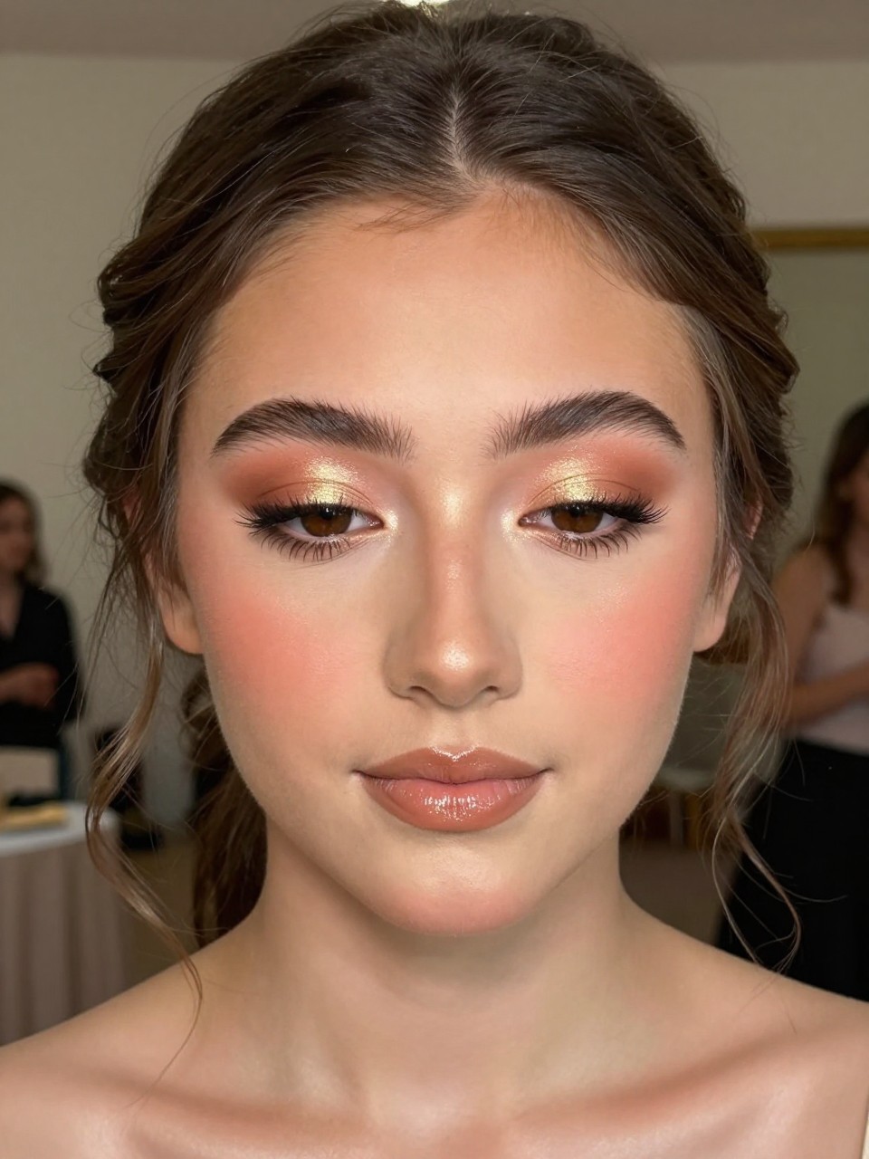 Super realistic close-up beauty portrait of a teenage woman with brown eyes wearing cute prom makeup, featuring golden peach shimmer eyeshadow on the lids, warm tan crease blending, golden peach blush on the cheeks and temples, light gold highlight on the high points, nude peach-beige glossy lips, softly lifted lashes, polished brows, realistic skin texture with a radiant warm glow, shot from a front-facing beauty portrait, with flattering warm prom lighting that clearly reveals the monochrome peach-gold tones, glossy lips, and luminous skin, soft romantic updo with loose tendrils, blurred elegant ballroom background, feminine modern beauty photography, latest iPhone photo quality.
