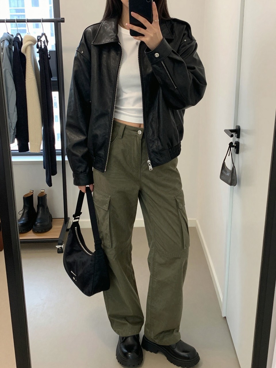 mirror selfie of a woman wearing the exact outfit described above, clearly showing a black relaxed-fit faux leather jacket, white fitted cropped tee, olive cargo pants with roomy side pockets, black chunky boots, and a small black nylon shoulder bag, holding a phone in front of a mirror, cropped at NECK so the face does not show, with a relaxed casual selfie pose, in a stylish city apartment, natural window light, including a clothing rack with garments behind and a handbag hanging on a door handle, latest iPhone photo quality.