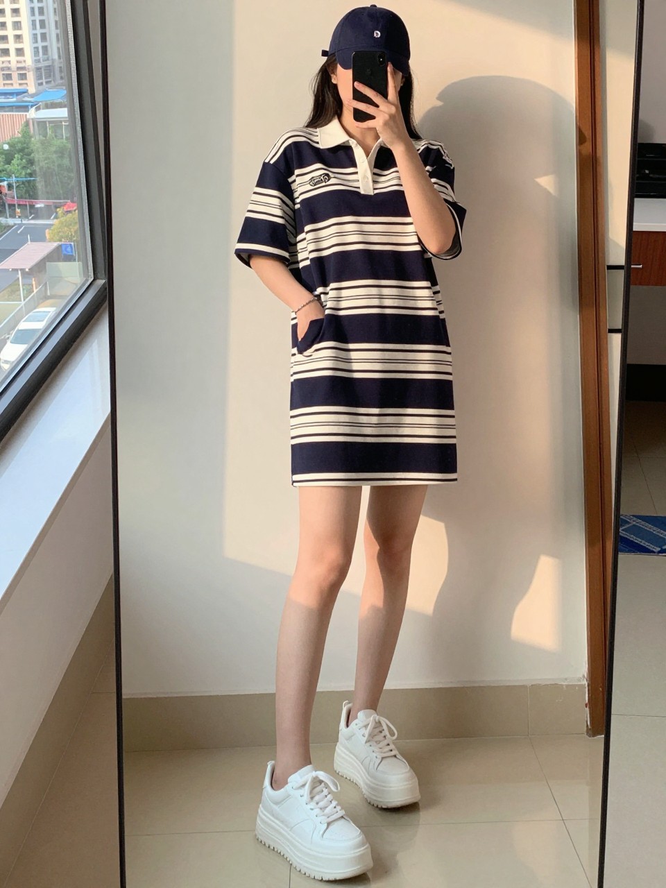 iPhone mirror selfie of a woman wearing a navy and white striped polo dress and chunky white platform sneakers with a matching navy baseball cap, posed with one hand in her pocket, cropped at NECK so face doesn't show, in a city apartment, evening golden hour, iPhone photo quality.