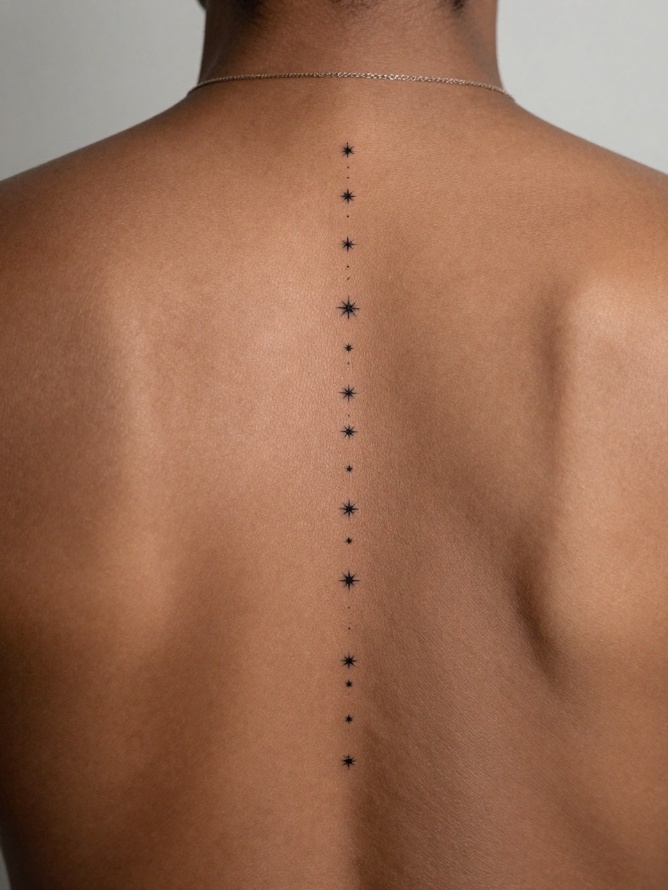 extreme close-up of a minimal star trail spine tattoo on deep brown skin, tiny black ink stars aligned down the center of the back, crisp fine line work, minimalist elegant style, realistic skin texture, delicate necklace chain visible, soft minimal background, design-focused tattoo inspiration photography (no watermarks on images)