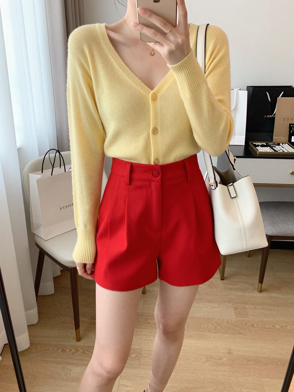 mirror selfie of a woman wearing a butter yellow knit cardigan with a soft fitted shape, bright red high-waisted tailored shorts, nude strappy sandals, a white structured shoulder bag, and a delicate gold layered necklace, holding a phone in front of a mirror, cropped at NECK so the face does not show, with a relaxed casual selfie pose, in a feminine-themed bedroom, natural window light, including shopping bags beside a chair and a jewelry tray on the vanity, latest iPhone photo quality. (no watermarks on images)