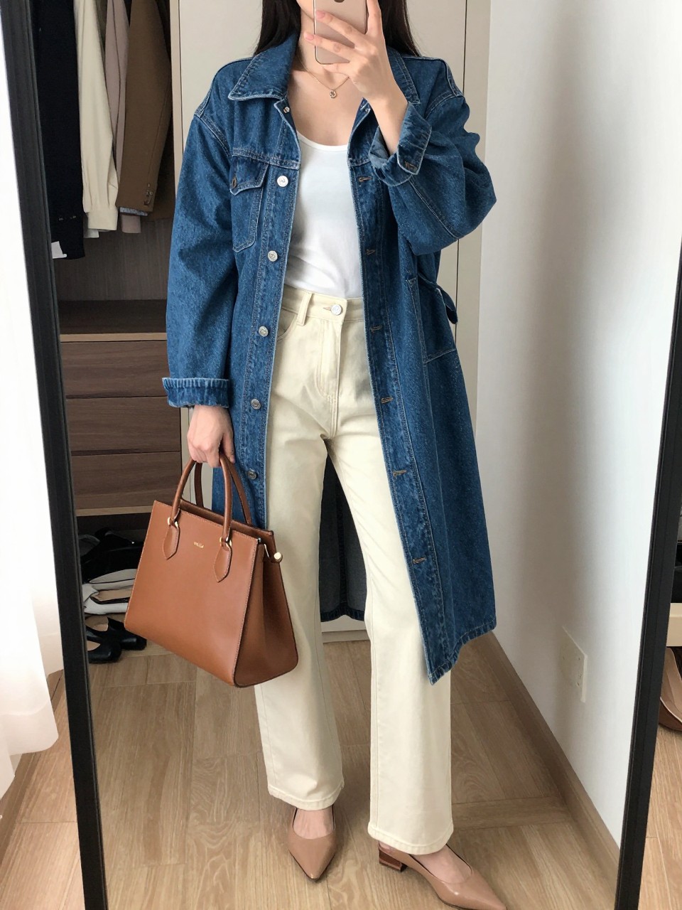 mirror selfie of a woman wearing the exact outfit described above, clearly showing a longline medium blue denim jean jacket, white fitted scoop-neck tank, cream wide-leg rigid denim jeans, nude pointed flats, and a brown structured top-handle bag, holding a phone in front of a mirror, cropped at NECK so the face does not show, with a relaxed casual selfie pose, in a walk-in closet mirror, natural window light, including an open closet with clothes visible and pair of heels on the floor nearby, latest iPhone photo quality.